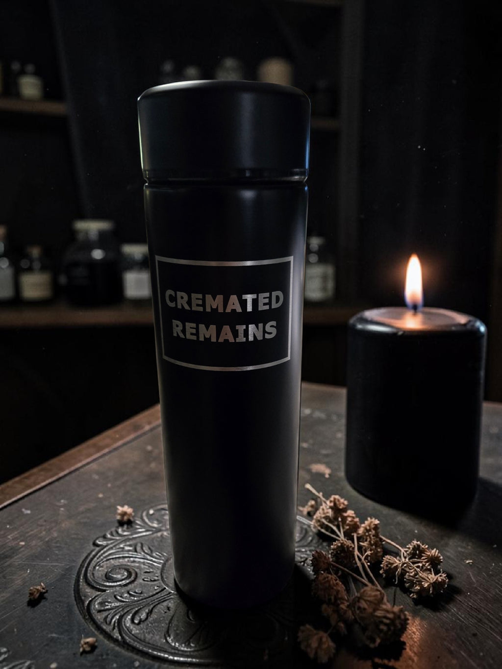 Black container labeled 'Cremated Remains' on a dark surface with a candle in the background.