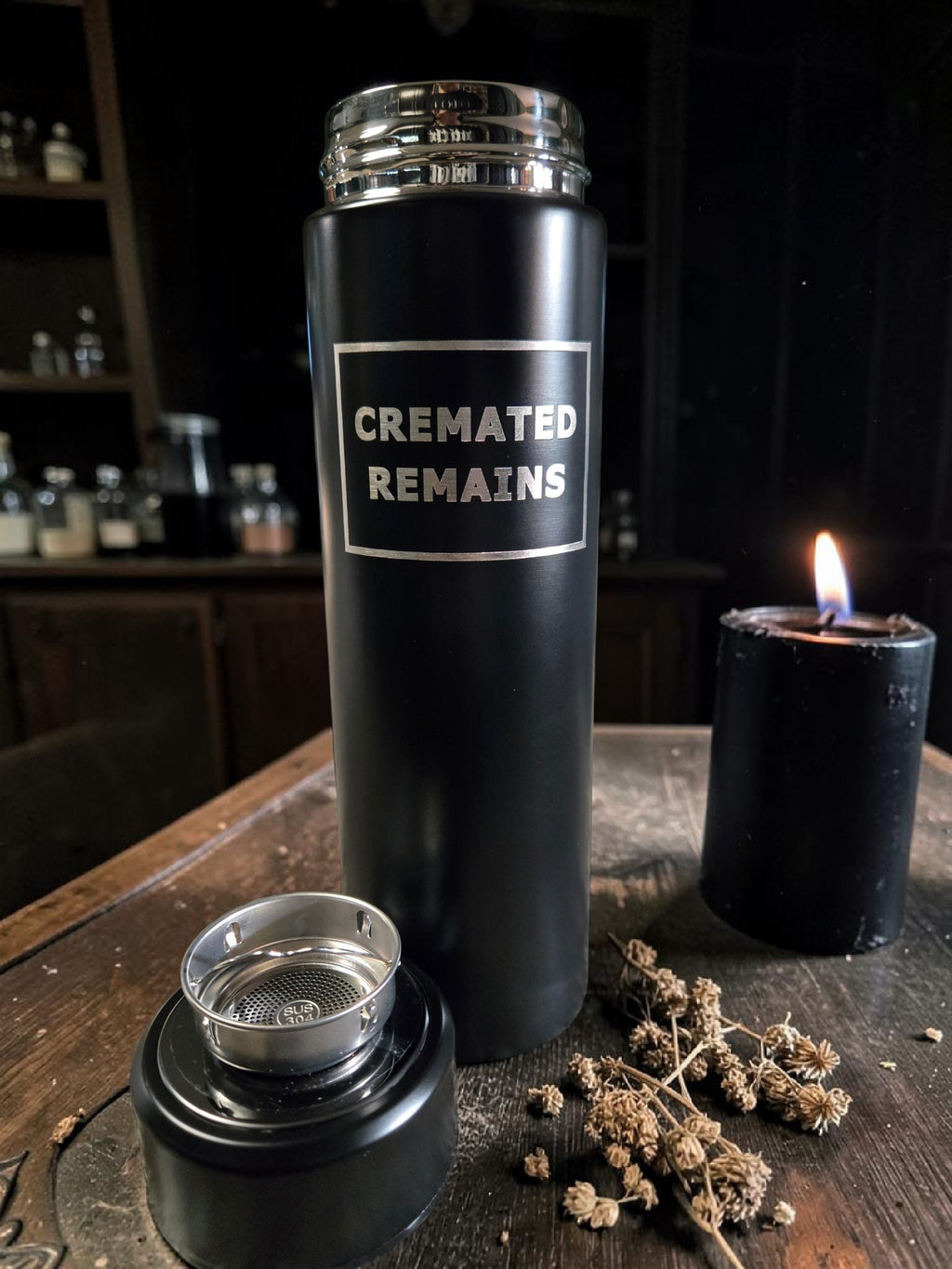 Black thermos labeled 'Cremated Remains' on a wooden surface with a candle and dried flowers.