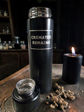 Black thermos labeled 'Cremated Remains' on a wooden surface with a candle and dried flowers.