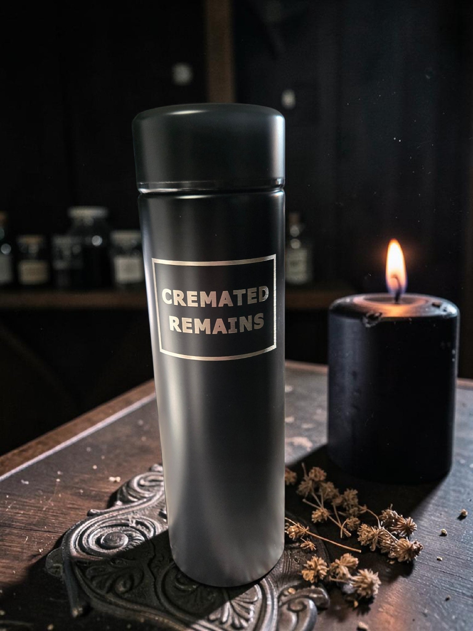 Black urn labeled 'Cremated Remains' on a dark surface with a candle and decorative elements.