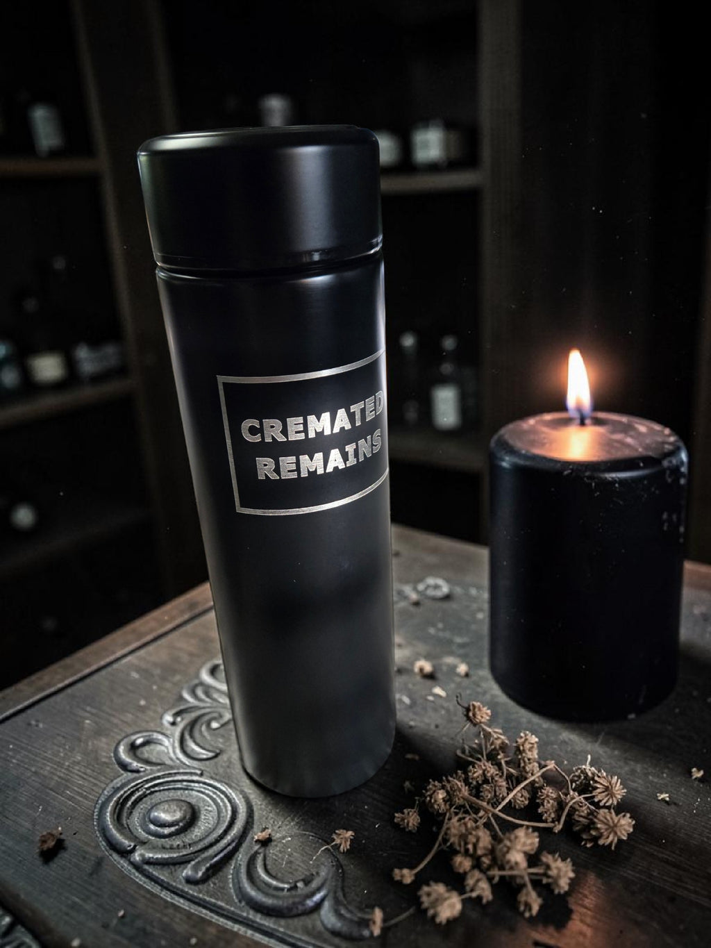 Black travel mug with 'CREMATED REMAINS' label next to a candle on a dark surface.