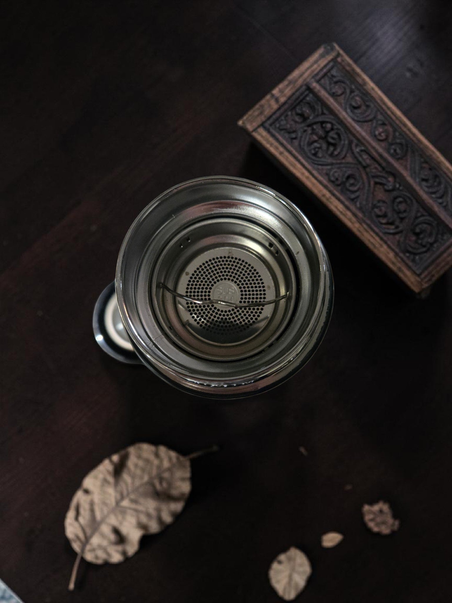 Metallic tea strainer on a dark wooden surface with decorative box and leaves.