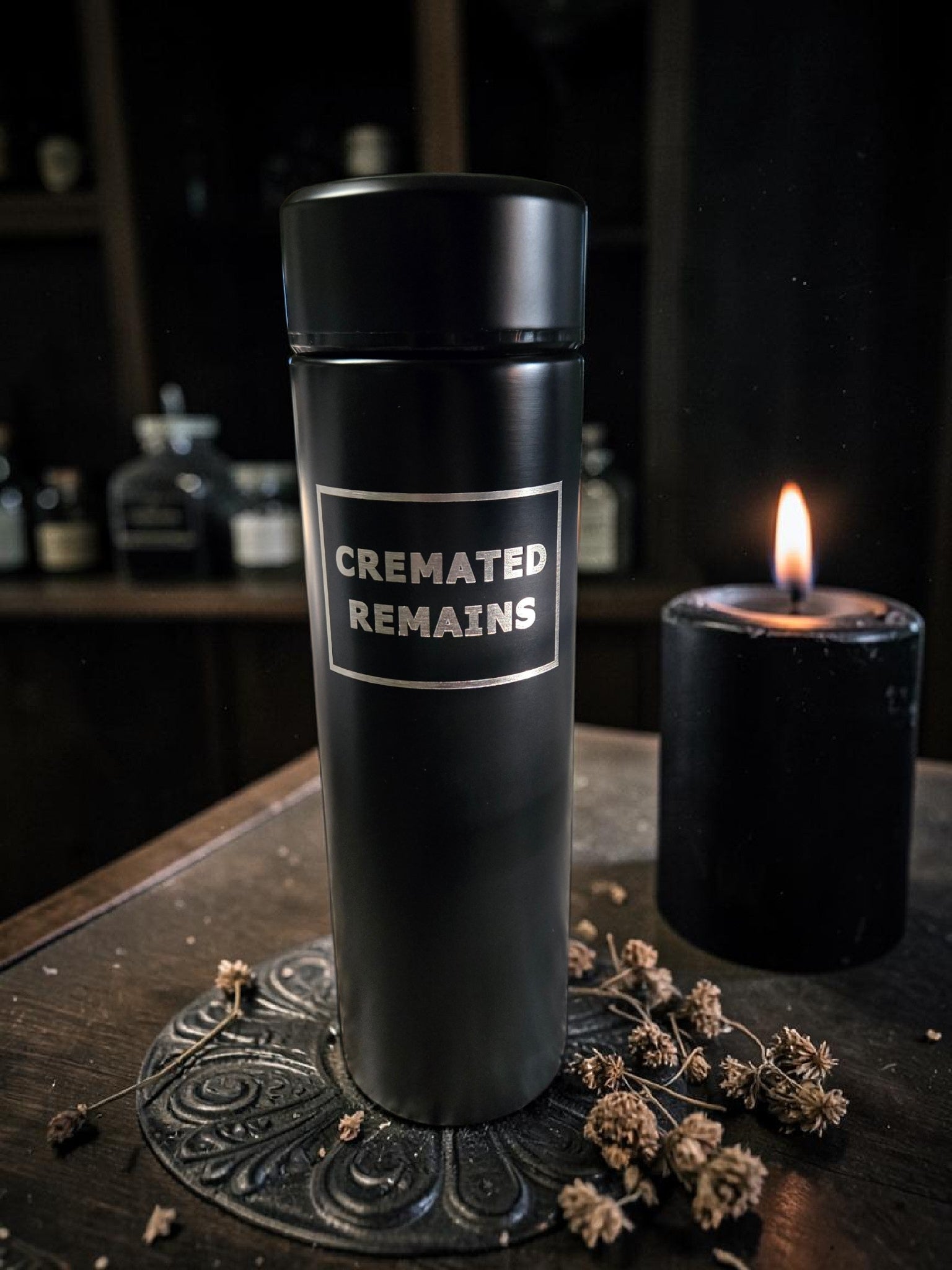 Black urn labeled 'CREMATED REMAINS' on a dark surface with a candle and dried flowers.